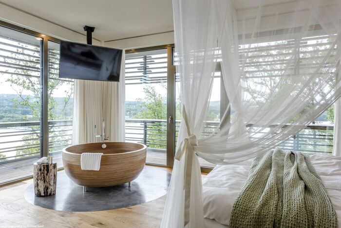 Runde Holzbadewanne, die im Doppelzimmer an der Fensterfront steht. Eine Doppelbett mit weißen Chiffonvorhängen.