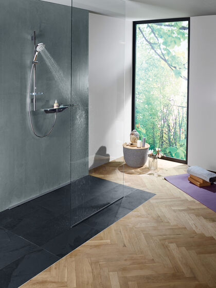 Helles Badezimmer mit Holzfußboden und Blick ins Grüne. Links eine Bodenebene Dusche mit schwarz gefliestem Boden und Grauen Wänden. In der Dusche ist die Duscharmatur Wellfit von HANSA installiert.