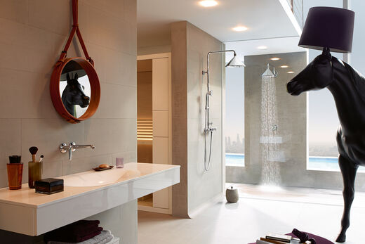 Badezimmer, in dem eine Axor ShowerProducts by Front Dusche installiert ist. Links befindet sich ein Waschtisch mit Waschbecken und Spiegel sowie Dekoelementen. Rechts im Hintergrund befindet sich eine Außendusche. Rechts vorne ragt ein Deko-Pferdekopf ins Bild hinein.