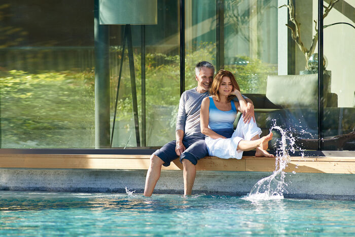 Lachendes Paar am Hotelpool hält die Beine ins Wasser.