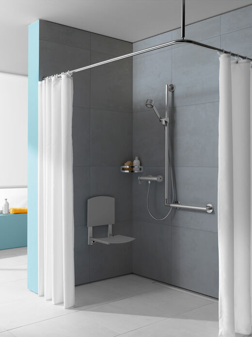 KEUCO Plan Care Echte Komfortdusche von KEUCO Plan Care mit Duschklappsitz in grau, stabilen Brausestangen-Haltegriffen in Chrom matt, Handbrause und Eckablage. Situation mit weißem Duschvorhang vor grauer Wand.
