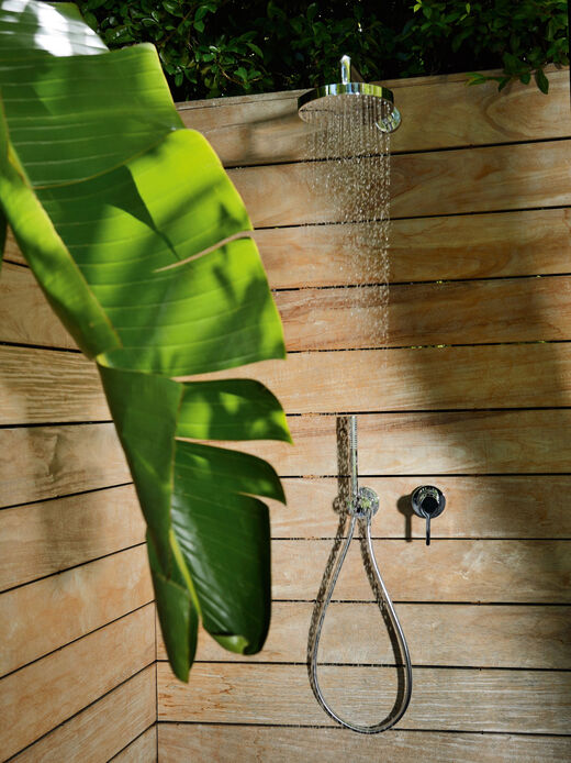 Keuco Ixmo Badarmatur, die in einer Aussendusche mit Holzwänden installiert ist. Aus der Kopfbrause fliest Wasser, die Handbrause ist an der Wand befestigt. Im Vordergrund befindet sich ein großes Blatt eines Bananenbaums, im Hintergrund sind grüne Blätter sichtbar.