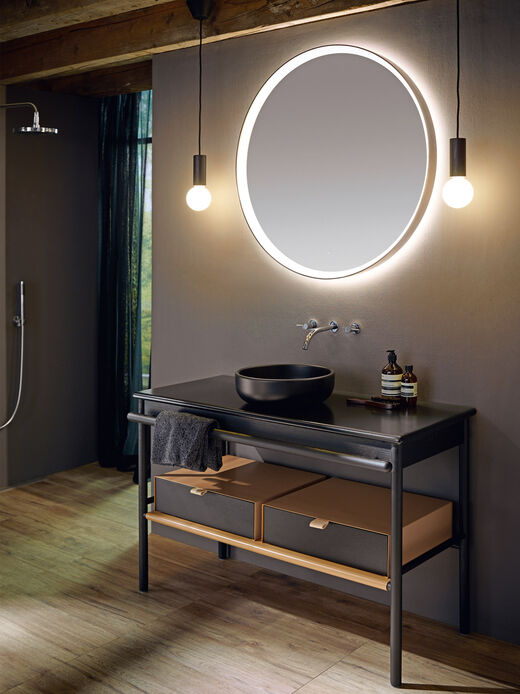 Runder beleuchteter Spiegel über modernem Waschplatz mit einem schwarzen Aufsatzwaschbecken und Holzmöbel.