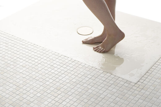 Beige Bette AntiRutsch Duschfläche, die in einem mit kleine Quadraten gefließten Boden eingelassen ist. Zwei weibliche Füße laufen gerade auf die Duschfläche.