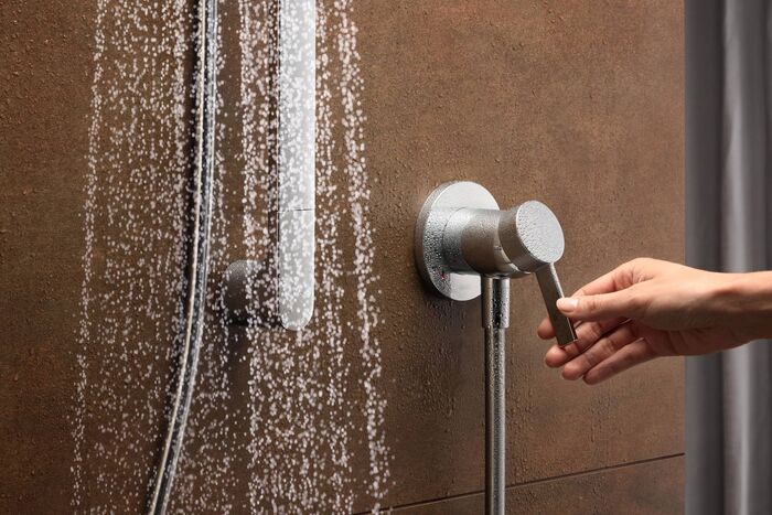 An einer braun - goldenen Wand ist eine Keuco Ixmo Armatur installiert, die gerade von einer weiblichen Hand betätigt wird. Links ist ein Wasserstrahl, der vermeintlich aus einer Duschbrause kommt, zu erkennen.