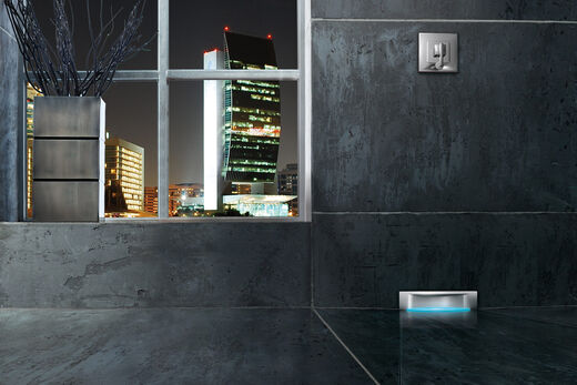 Grau geflieste Badezimmerwand, an der im rechten Teil des Bildes ein beleuchteter Wandablauf installiert ist. Links befindet sich ein Fenster mit Blick in eine Großstadt mit Wolkenkratzern.
