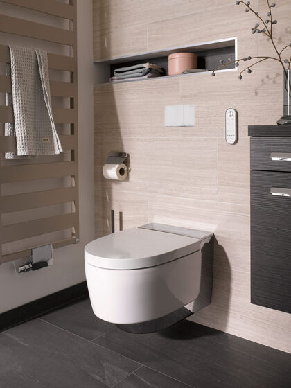 Dusch-WC-Szene von Geberit: Das Modell AquaClean Mera wird an einer hellen Holzwand von einem Badheizkörper und einem Badmöbel flankiert. 