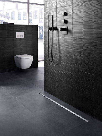 In grau-schwarz gehaltener Raum, in dem an einer gefliesten Wand eine Duscharmatur befestigt ist. Auf dem Boden befindet sich eine Geberit CleanLine Duschrinne. Links im Bild ist eine Kloschüssel befestigt