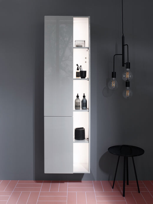 Ein grauer Hochschrank der Serie Badu, wandhängend, im Badezimmer mit seitlichen Staufächern für Shampoos und Pflegeprodukte. 