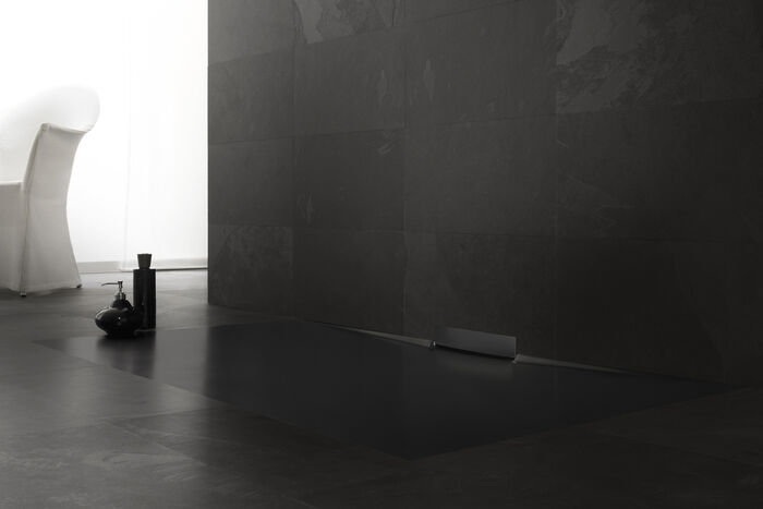 Schwarze Kaldewei Xetis Duschfläche, die in einem grauen Boden eingelassen ist, dahinter befindet sich eine grau geflieste Wand. Links stehen Hygieneartikel auf dem Boden, im Hintergrund ist eine Sitzgelegenheit zu erkennen.