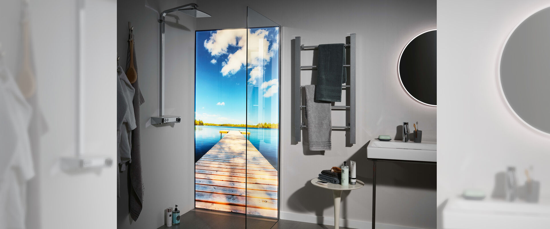 Dusche, in der eine Premium LED Duschwand von Spritz installiert ist, auf der ein Steg, der aufs Meer hinaufführt abgebildet ist. Die Dusche befindet sich in einem ansonsten grau gehaltenem Badezimmer. Links von der Dusche ist ein Handtuchhalter, darunter ein Tisch als Ablagemöglichkeit sowie ein Waschbecken mit Spiegel zu erkennen.
