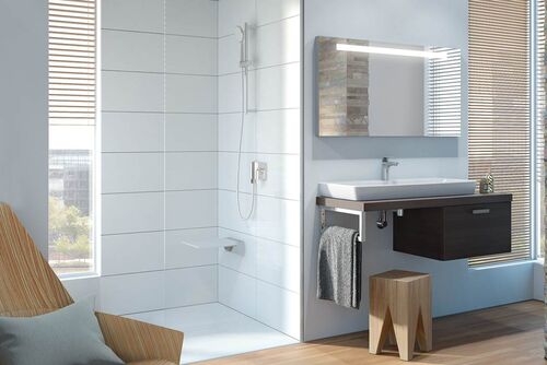 Komfortbad Conforma von VitrA lässt viel Raum zum Entspannen. Große, offene Dusche vor einem Fenster, Sessel, wohnlicher Waschplatz aus Unterschrank und Waschtisch sowie Sitzhocker kombiniert mit einem beleuchteten Spiegelschrank.