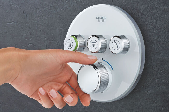 GROHE Smart Control Duscharmatur in kreisrunder Form, die an einer grauen Wand installiert ist. Auf der Oberfläche befinden sich insgesamt 4 verschiedene Regler, der unterste wird gerade von einer Hand betätigt.