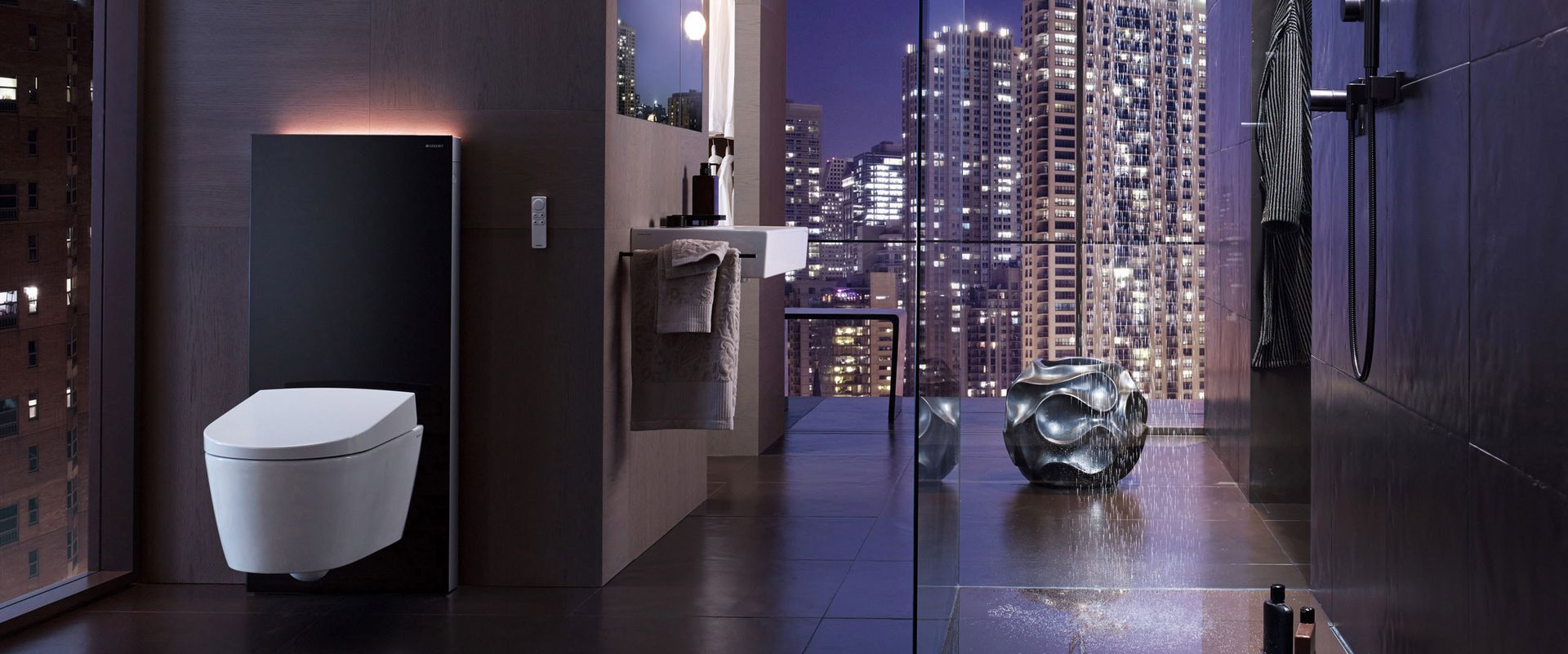 Beeindruckendes Design-Badezimmer von Geberit mit Aussicht auf eine urbane Skyline. Punktuell beleuchtete Nachtszene mit Dusch-WC AquaClean auf Monolith-Modul, daneben Waschtisch und Dusche mit angeschaltetem Wasser.
