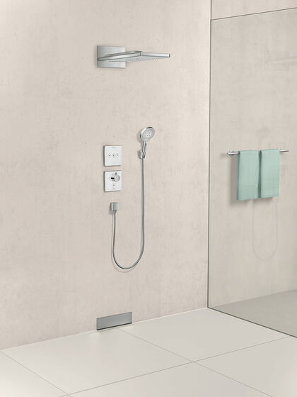 Beige gehaltene Dusche, in der eine rainmakerSelect Kopfbrause von Hansgrohe installiert ist. Hinter der gläsernen Duschwand hängen zwei turkiese Handtücher an einer Armatur.