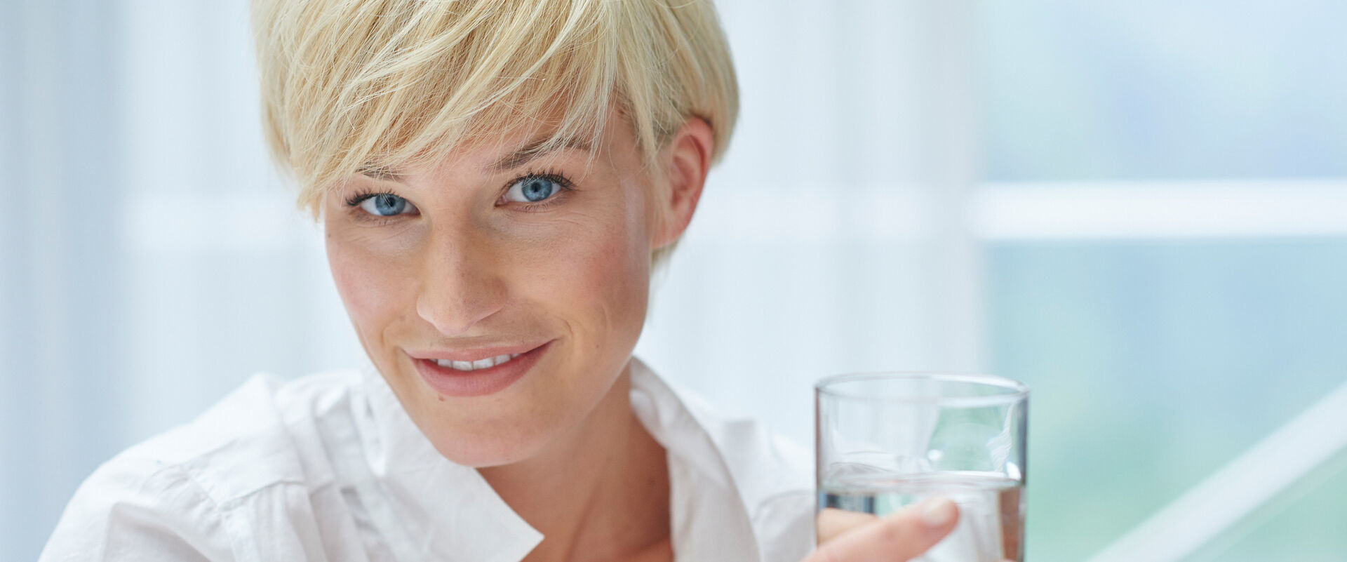 Frau mit Wasserglas. Blick frontal, kurze blonde Haare. hintergrund hellblau.