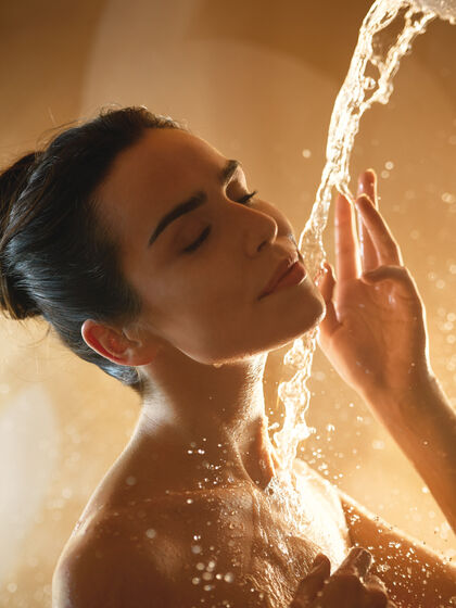Frau duscht in goldenem Licht. ein Wasserstrahl läuft ihr über den Oberkörper.