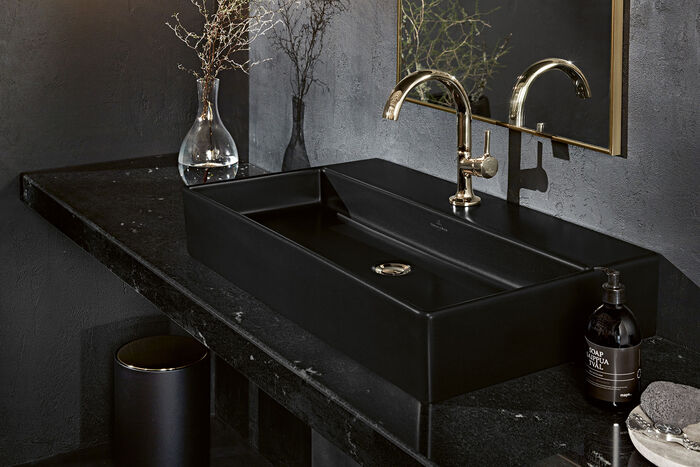 Premium-Waschplatz von Villeroy & Boch mit außergewöhnlichem, schwarzem Keramik-Aufsatzwaschbecken aus der Serie Memento 2.0, schwarzer Konsole, Vase und goldfarbenem Wasserhahn.