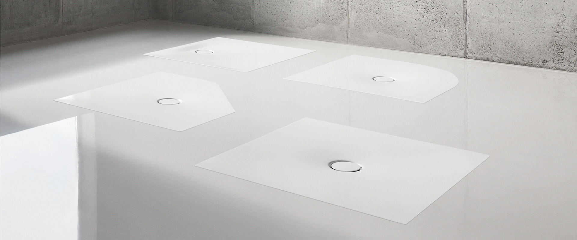 Grauer Boden, auf dem vier weiße BetteFloor Duschflächen von Bette in unterschiedlichen Formen eingelassen sind.
