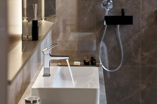 hansgrohe Metropol Eckiges Waschbecken, an dem die Metropol - Badarmatur der Marke hansgrohe installiert wurde. Wasser läuft durch den Wasserhahn, links ist ein beleuchteter Badezimmerschrank aufgehängt. Im Hintergrund sind Elemente einer Dusche zu sehen.