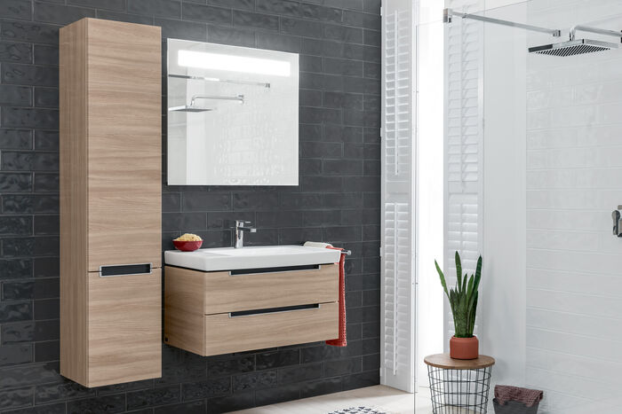 Hoher Wandschrank in hellem Holz, daneben ein Waschbecken in einem Badmöbel integriert. Darüber ein Spiegel.