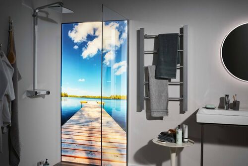 Dusche, in der eine Premium LED Duschwand von Spritz installiert ist, auf der ein Steg, der aufs Meer hinaufführt abgebildet ist. Die Dusche befindet sich in einem ansonsten grau gehaltenem Badezimmer. Links von der Dusche ist ein Handtuchhalter, darunter ein Tisch als Ablagemöglichkeit sowie ein Waschbecken mit Spiegel zu erkennen.