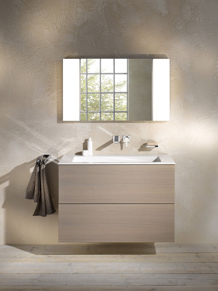 Keuco Edition 11 Waschbecken Edition 11 von Keuco, das an einer beigen Wand installiert ist. Auf dem Waschbecken stehen Hygieneartikel, darüber hängt ein Spiegel.