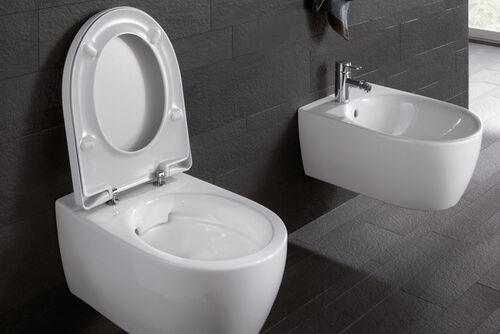 Kombination vor schwarzer Wand aus Bidet und dem spülrandlosem WC Rimfree® von Geberit mit geöffnetem Deckel während des Spülvorgangs.
