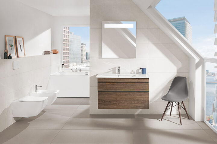 Helles Badezimmer mit frontalem Blick auf den Waschplatz mit wandhängendem Unterschrank aus Holz und Badspiegel. Seitlich Toilette und Bidet.