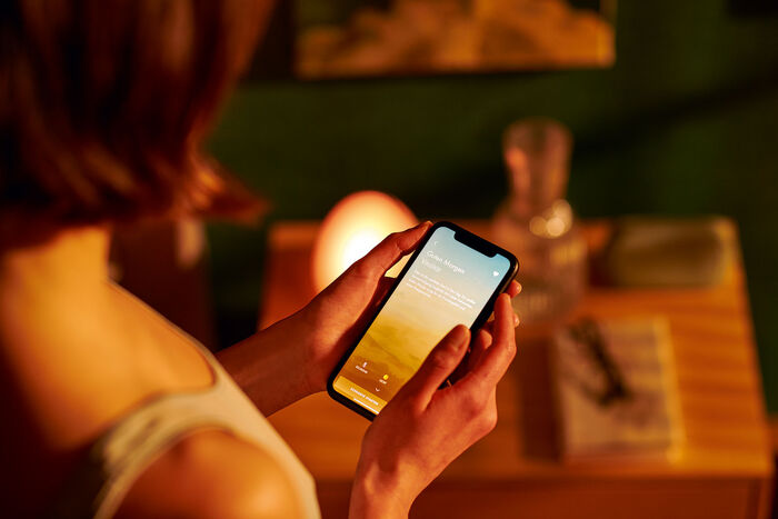 Mit der hansgrohe App lassen sich insgesamt 7 Duschereignisse auswählen, die dann über den Playbutton in der Dusche gestartet werden. Frau mit Smartphone in goldenem Licht.