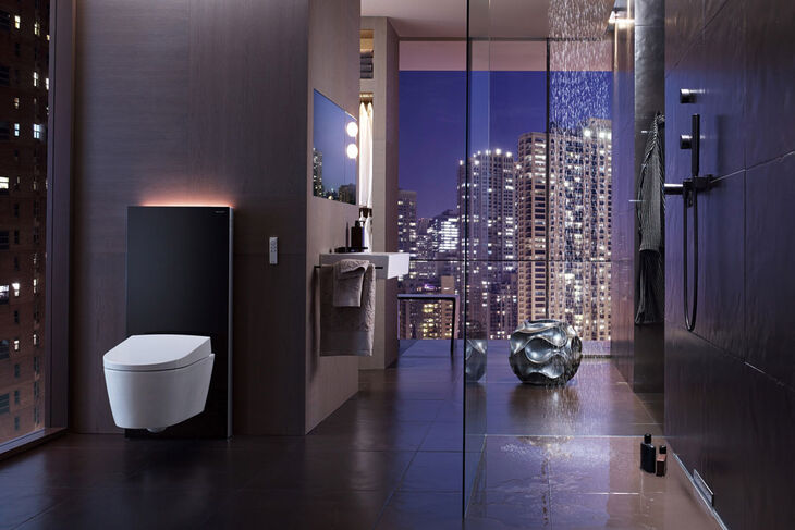 Stimmungsvolle Aufnahme bei Nacht eines Badezimmers mit der Skyline einer Großstadt. Links im Vordergrund thront ein Dusch-WC AquaClean von Geberit kombiniert mit dem Monolith von Geberit.