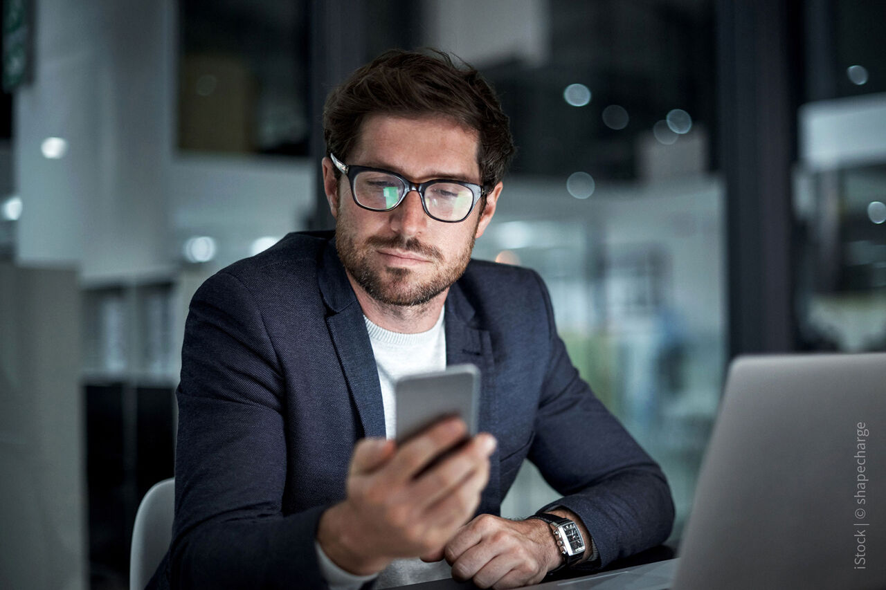 Businessmann mit Anzug und Brille, der auf sein Smartphone schaut und vor dem Laptop sitzt – SYR-Modelbild.