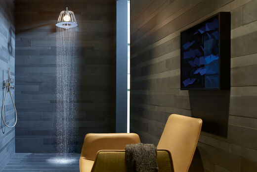 AXOR Lampshower by Nendo Im Vordergrund befindet sich rechts ein gelber Sessel, darüber ein Bild. Im Hintergrund ist eine AXOR Nendo Kopfbrause installiert, in der sich eine Lampe befindet. Aus der Kopfbrause fließt Wasser.