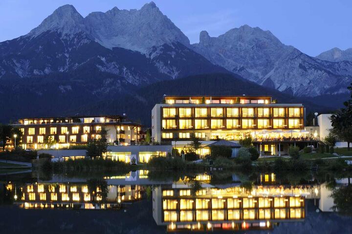 Hotel Ritzenhof in der Abendstimmung hell erleuchtet. Vor dem Haus der See, in dem sich das Hotel spiegelt und im Hintergrund die Berge.