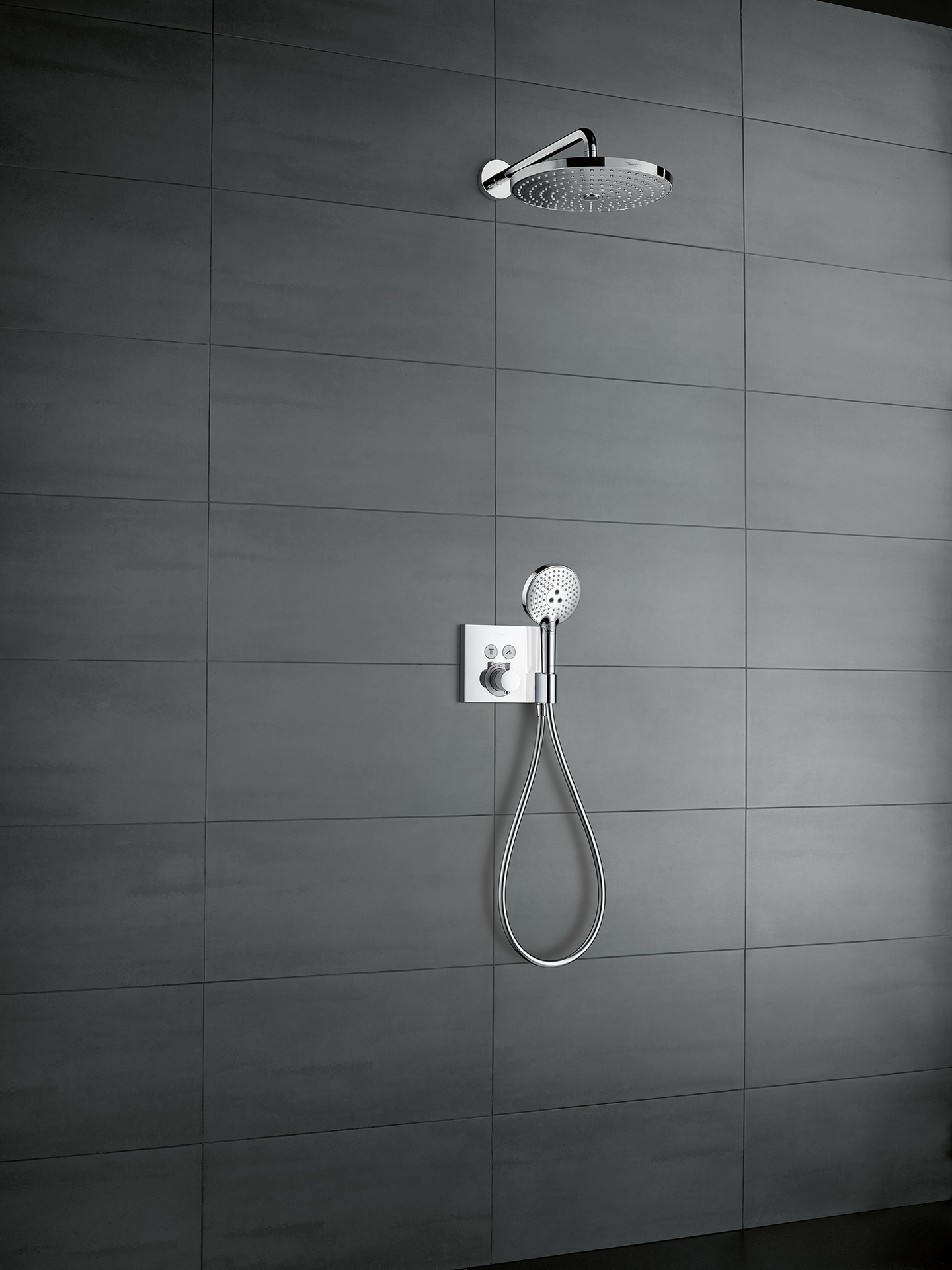 Grau geflieste Duschwand, an der ein rechteckiges hansgrohe ShowerSelect Thermostat befestigt ist. An diesem Thermostat befindet sich außerdem eine Handbrause mit Wasserschlauch. Darüber befindet sich eine Kopfbrause.