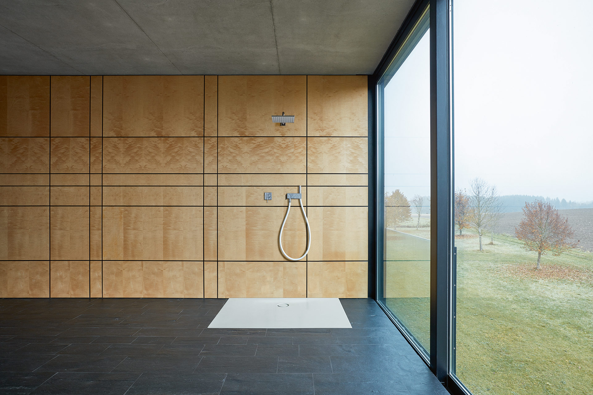 Heller Raum, auf der rechten Seite Fenster, die von Decke zu Boden reichen mit Blick ins Grüne. Schwarzer Boden, Wand, mit Holz verkleidet. An der Wand ist eine Dusche installiert, für die eine bodenebene Duschwanne BetteFloor von Bette montiert ist.