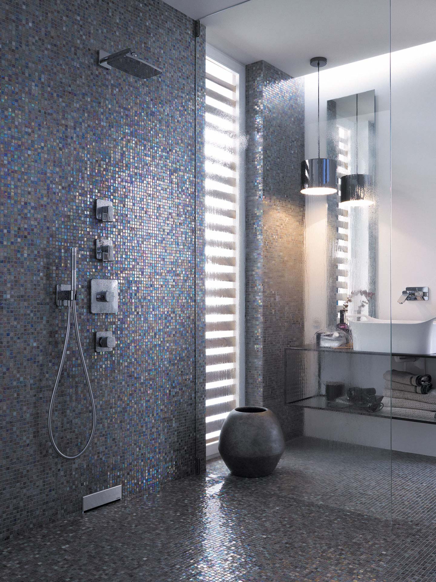 Badezimmer, das mit kleinen quadratischen Fliesen gefliest ist. Links im Vordergrund befindet sich eine Dusche, in der ein Geberit Wandablauf installiert ist. Rechts befindet sich ein Waschbecken mit Unterschrank, in dem sich Handtücher befinden.